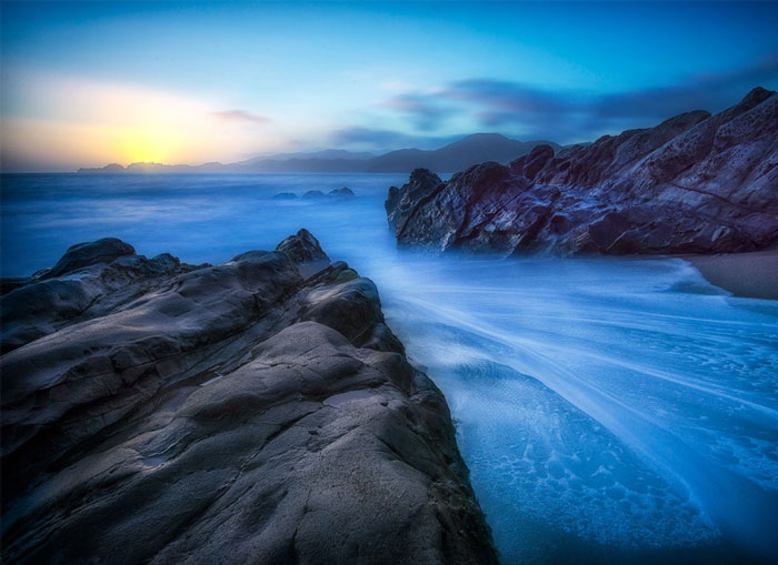 An image of rocky ridges with water and sun in the background