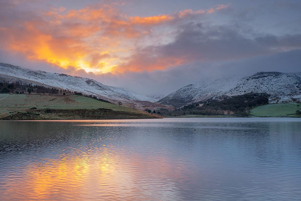 An image of snowy mountain with water in the foreground and sun rays in the background