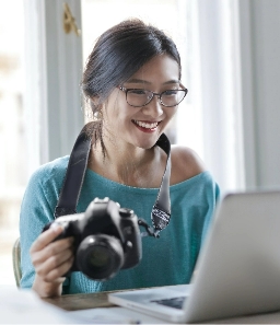 An image of a girl with glasses holding a camera in her hand and looking at the laptop