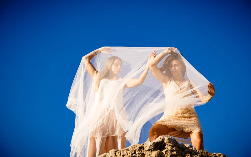 Two people holding sheer white fabric against a blue sky during an outdoor photo shoot