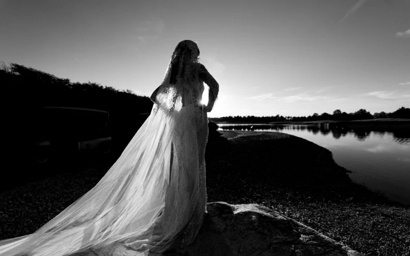 Black and white figure photo of a bride in a veil and flowing dress beside a lake