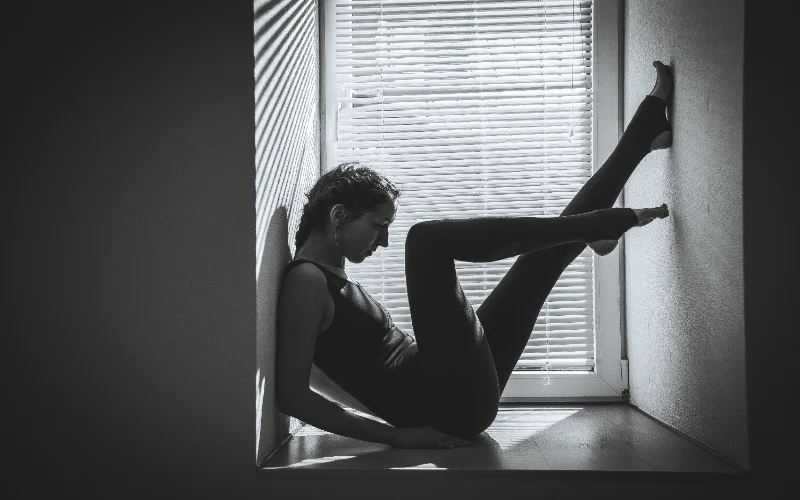 Black and white figure photography of a woman stretching against a window with blinds