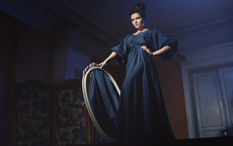 Woman in a dark blue gown holding a large hoop in a dimly lit vintage-style room for a artistic fashion photography