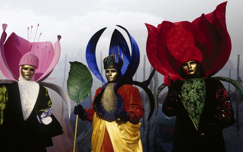 Models in elaborate carnival-style costumes and oversized headpieces against a stone backdrop