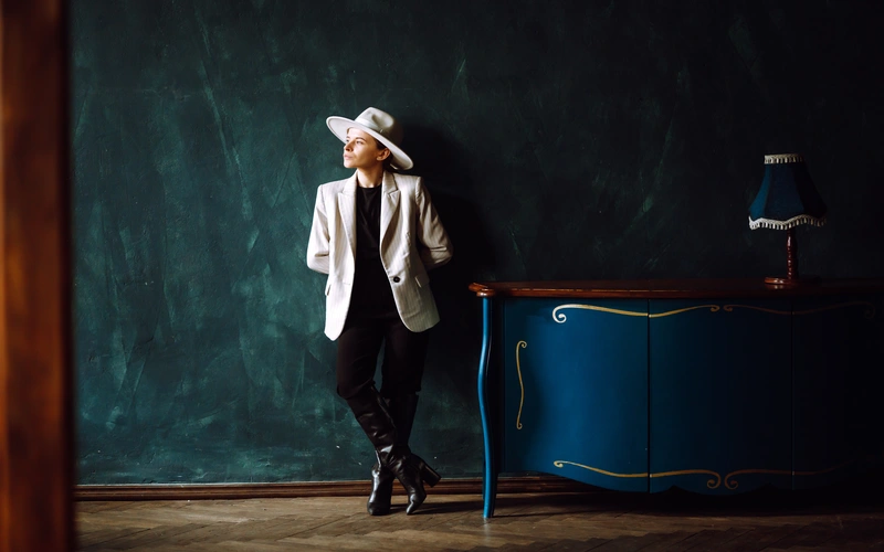 Woman in a white blazer and hat leaning against a vintage blue desk in a styled fashion portrait