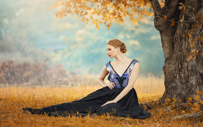 Woman in a deep blue gown sitting among golden autumn leaves beside a tree