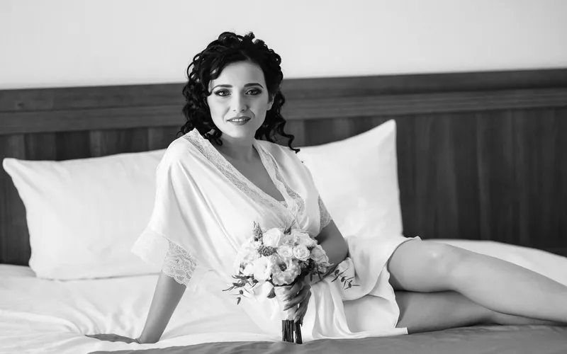 black and white Wedding boudoir portrait of a woman in a robe sitting on the corner of the bed