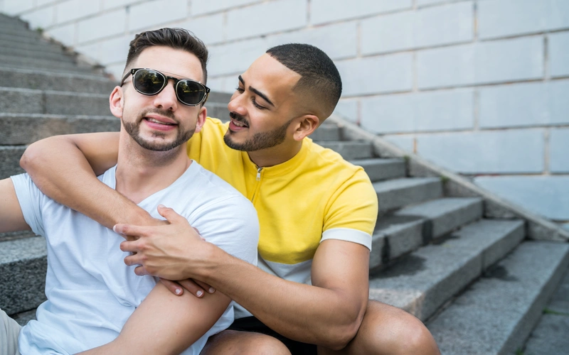 Gay couple sitting together on steps, one resting against the other in a natural, relaxed pose