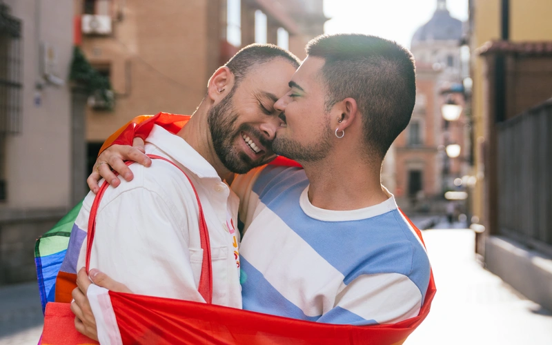Gay couple embracing closely on a city street, wrapped in a pride flag