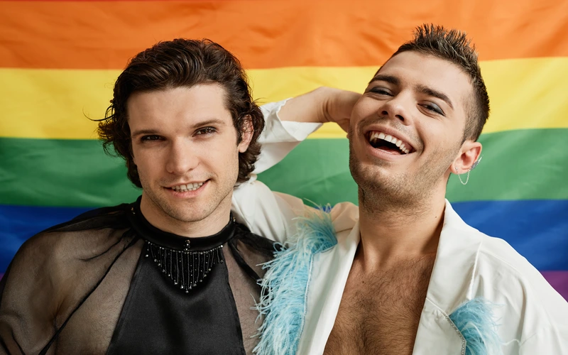 Two men smiling closely together against a rainbow pride flag backdrop