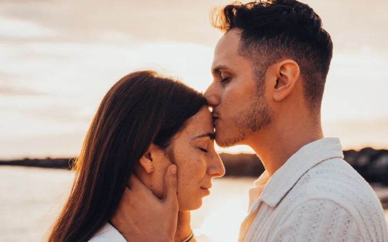 Romantic couple pose of a man kissing his partner's forehead at golden hour by the water