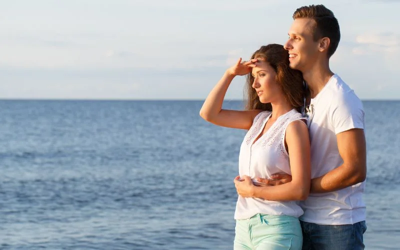 Couple gazing out at the ocean together, woman shielding her eyes from the sun