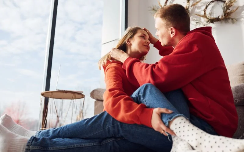 Intimate couple pose with the woman seated on her partner's lap by a sunlit window