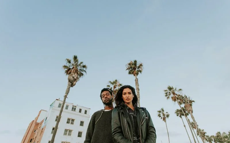 Wide-angle shot of a couple standing close together in front of palm trees and city buildings
