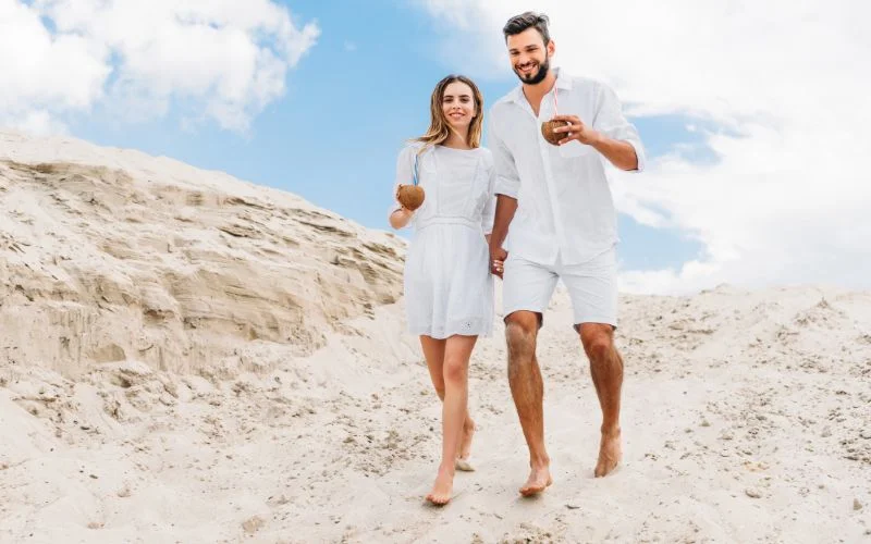 Couple in white outfits walking hand in hand along a sandy beach
