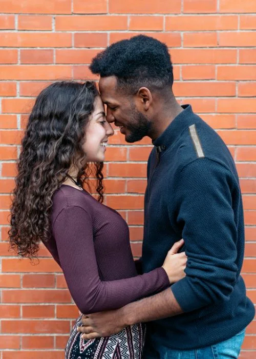 Couple standing forehead to forehead in an intimate embrace against a red brick wall