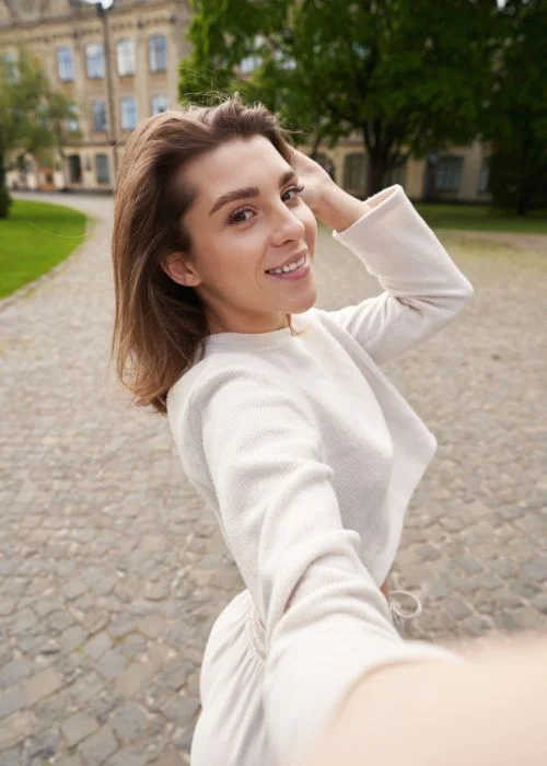Woman taking a selfie outdoors while gently touching her hair, demonstrating a natural and flattering selfie pose idea.