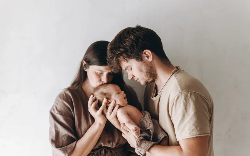 Newborn poses with parents showing a mother cradling and kissing her baby's forehead
