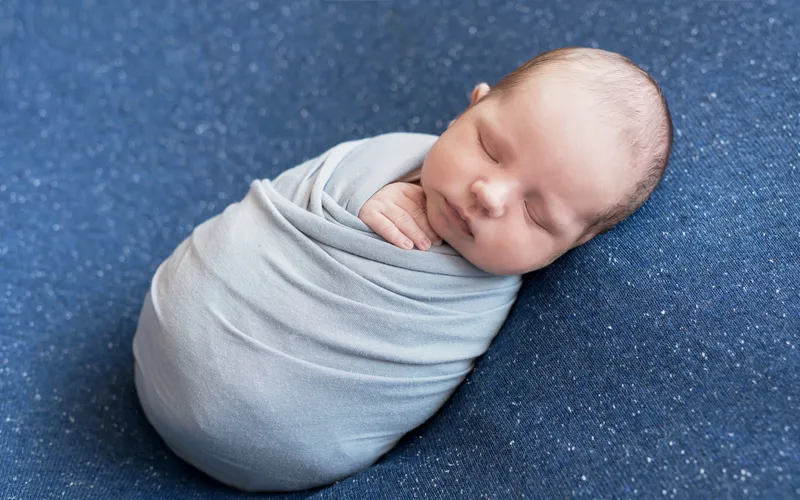 Newborn swaddled snugly in a white wrap with a floral bonnet, resting on cream fur