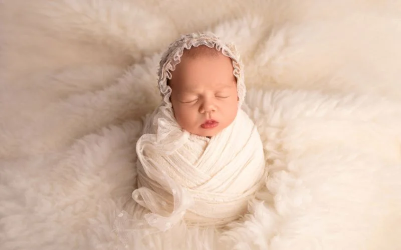 Newborn swaddled snugly in a white wrap with a floral bonnet, resting on cream fur