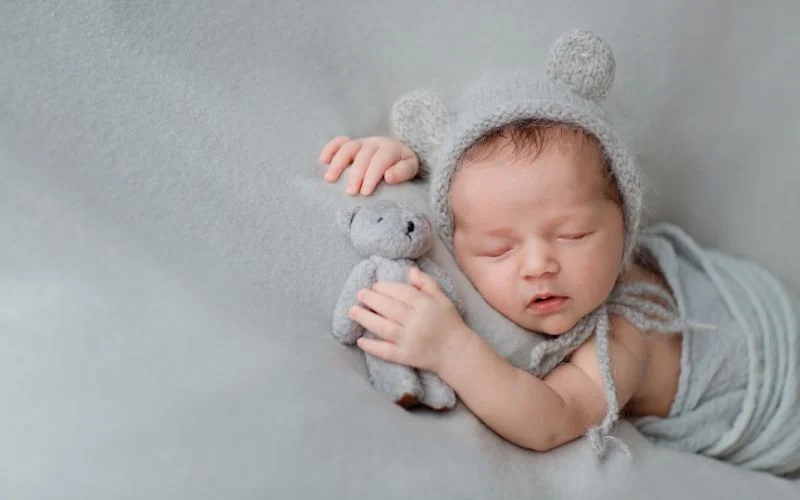 Sleeping newborn cuddled up with a soft knit mouse toy in a grey wrap