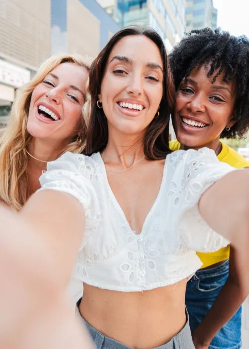 Three friends smiling and posing together for a cheerful group selfie in an urban setting, showcasing fun group selfie poses.