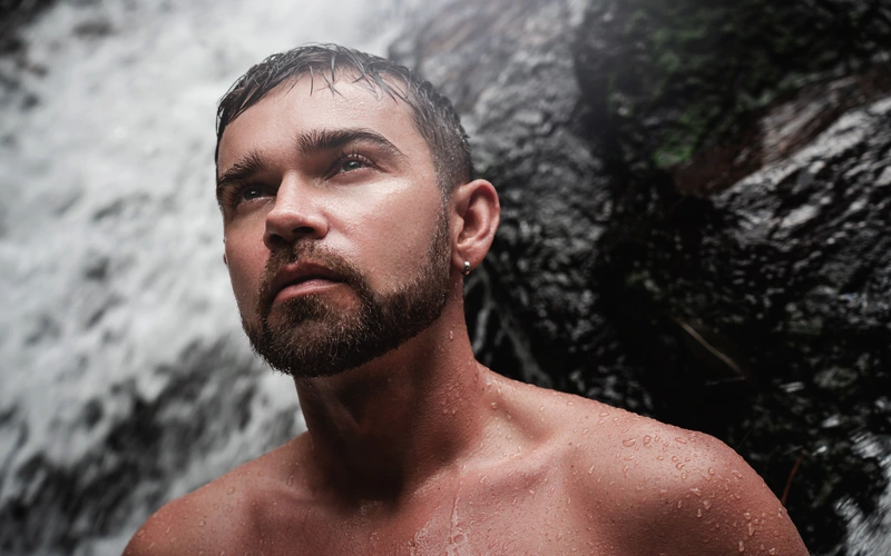 Man posing for boudoir photoshoot in nature with water droplets on his skin
