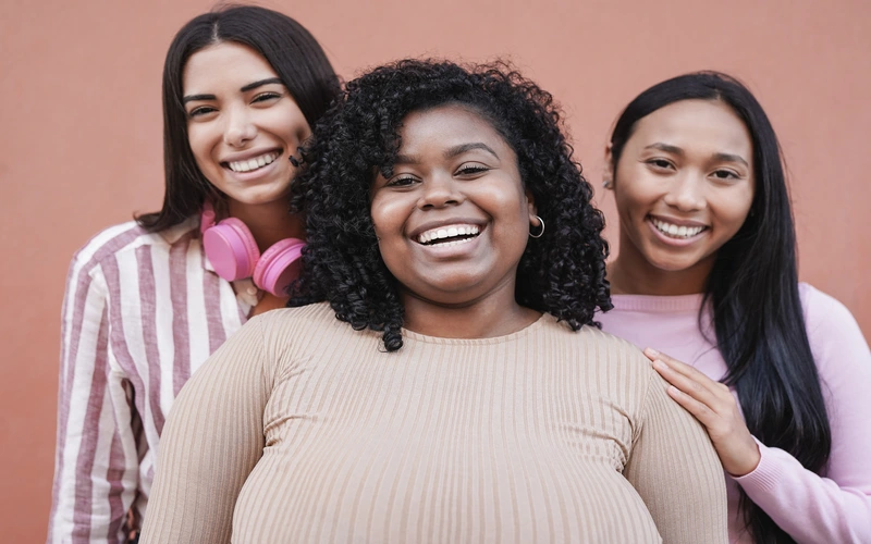 Three women smiling together against a pink background