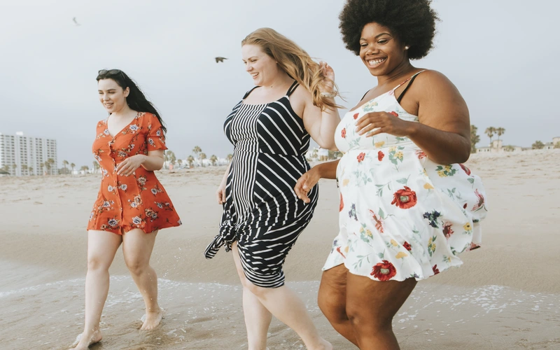 Three women of different body types walking together on a beach, laughing and relaxed