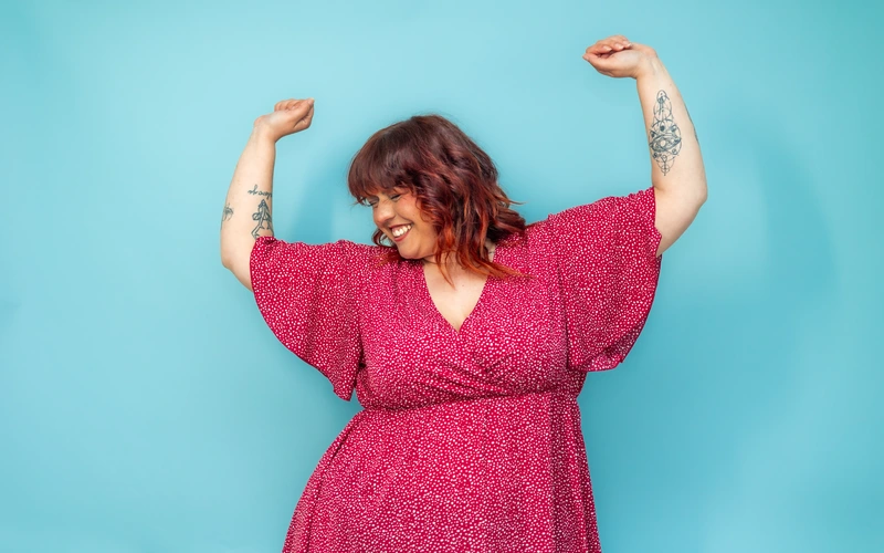 Woman with a curvy body type raising her arms confidently in a pink dress against a teal background