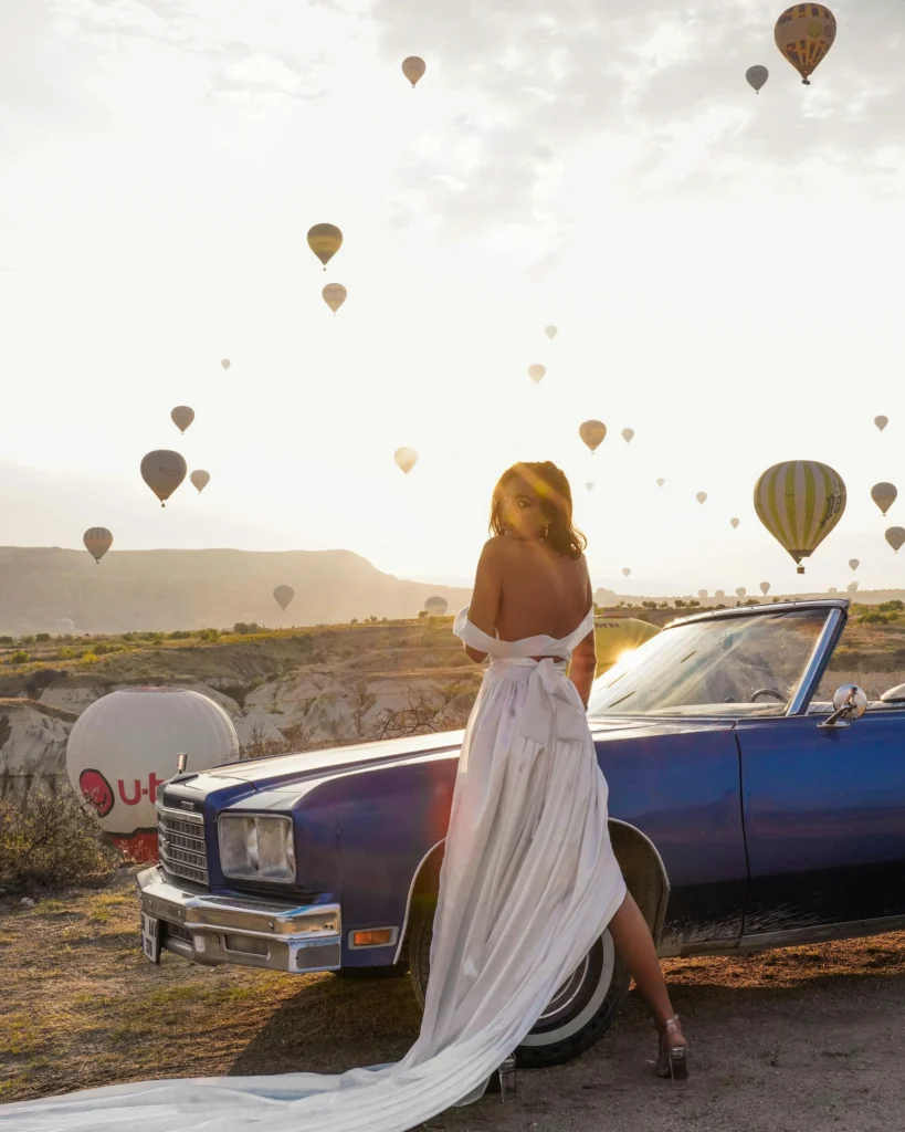 car photoshoot ideas - a model in a white gown posing next to a car during golden hour
