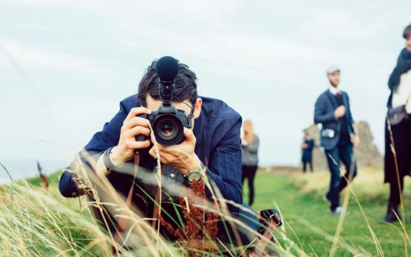 Photographer lying in tall grass adjusting camera settings outdoors, relevant to how is aperture measured