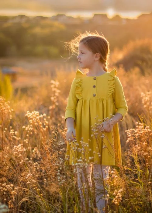 Young girl in a yellow dress standing in a sunlit field of tall grass during golden hour