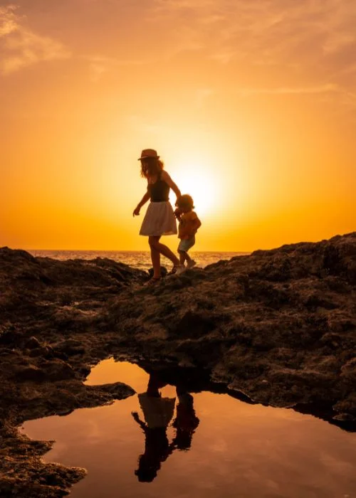 Silhouette of a mother and child walking along a rocky shoreline at sunset, golden hour photography reflected in a tide pool