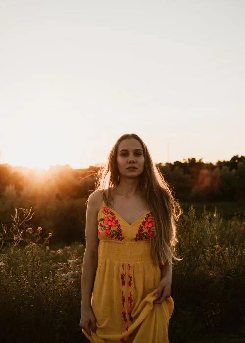 Woman in a yellow floral dress standing in a field, backlit by the setting sun with soft lens flare